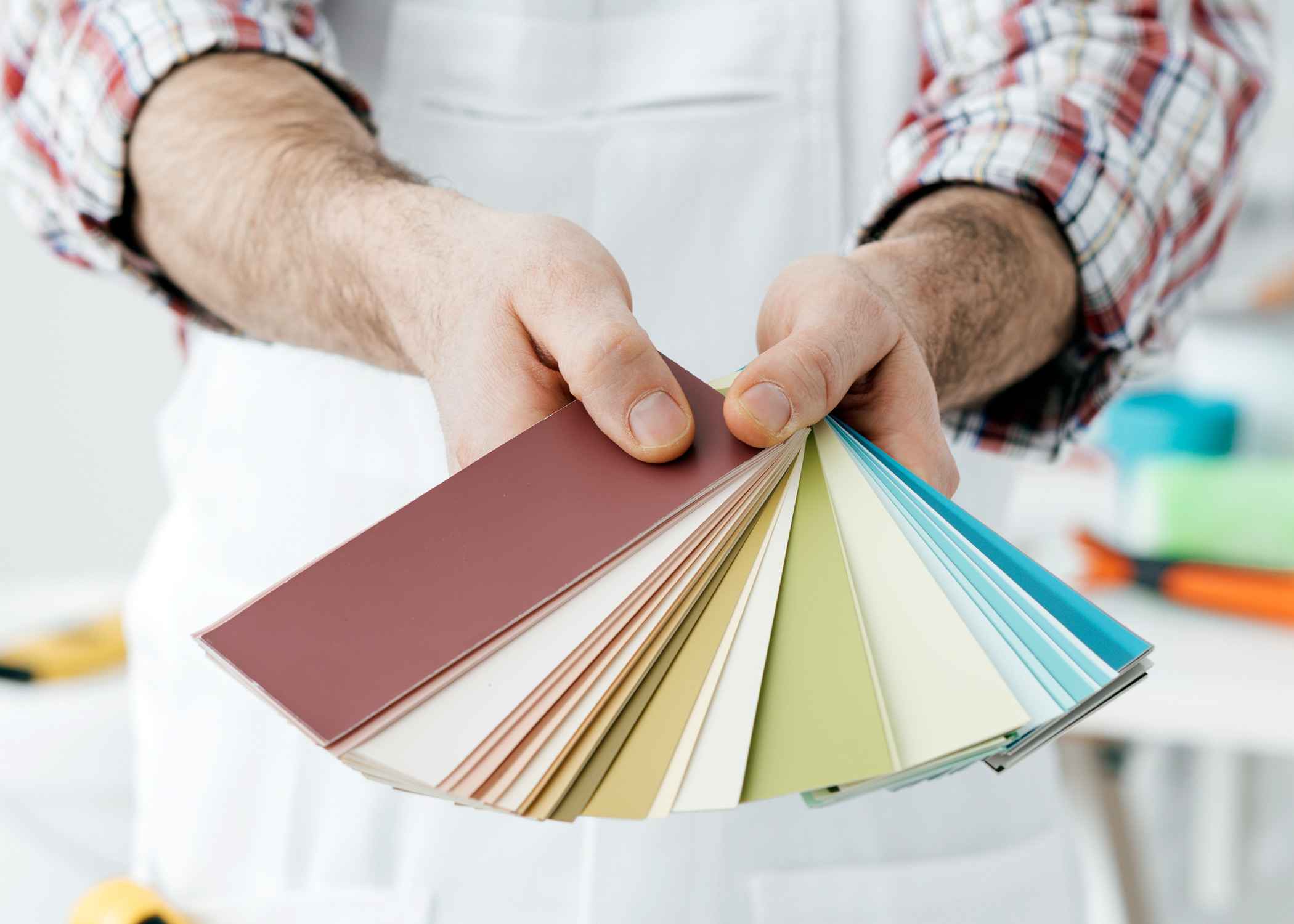 Person in a plaid shirt and white apron holding a fan of paint color samples, featuring shades from deep red to light blue and green.