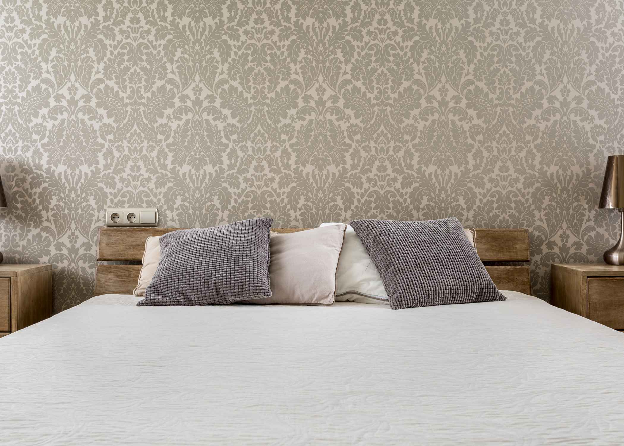 Bed with neutral pillows, wood nightstands, and beige patterned wallpaper in a cozy, symmetrical bedroom setup.