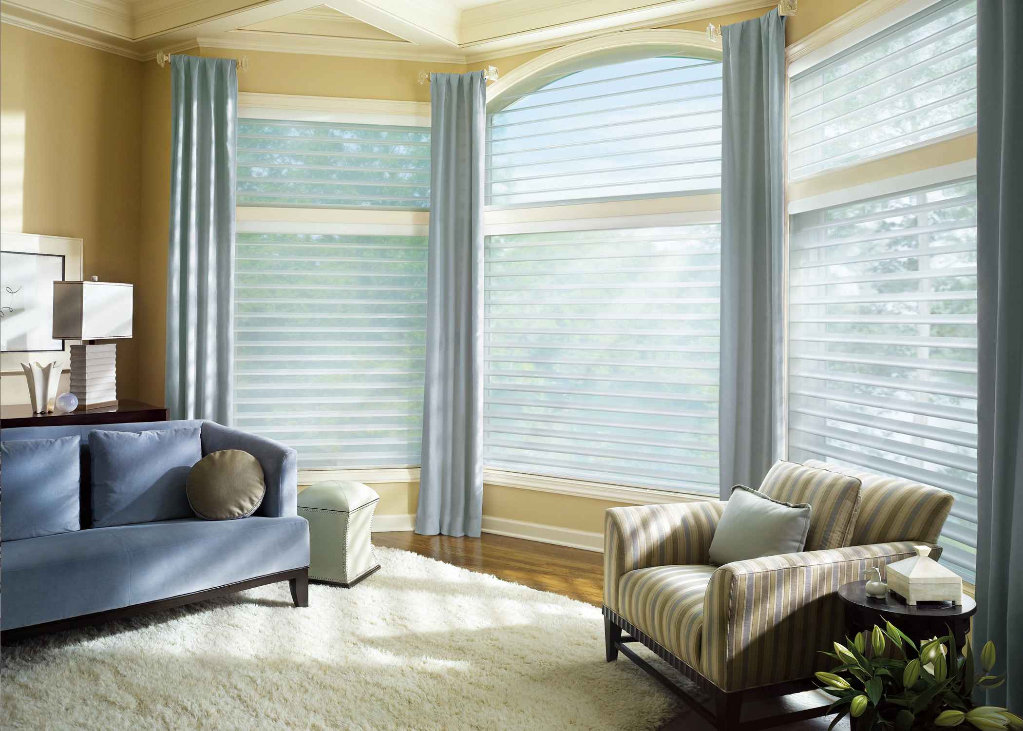 Living room with large windows covered by sheer layered blinds and light blue curtains, featuring a blue sofa, striped armchair, and cream rug.