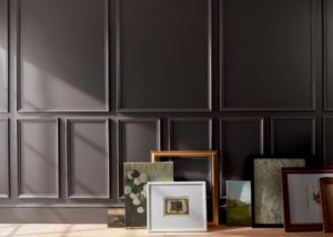 Artwork and frames arranged on a floor against a deep chocolate brown-painted wall with decorative molding.